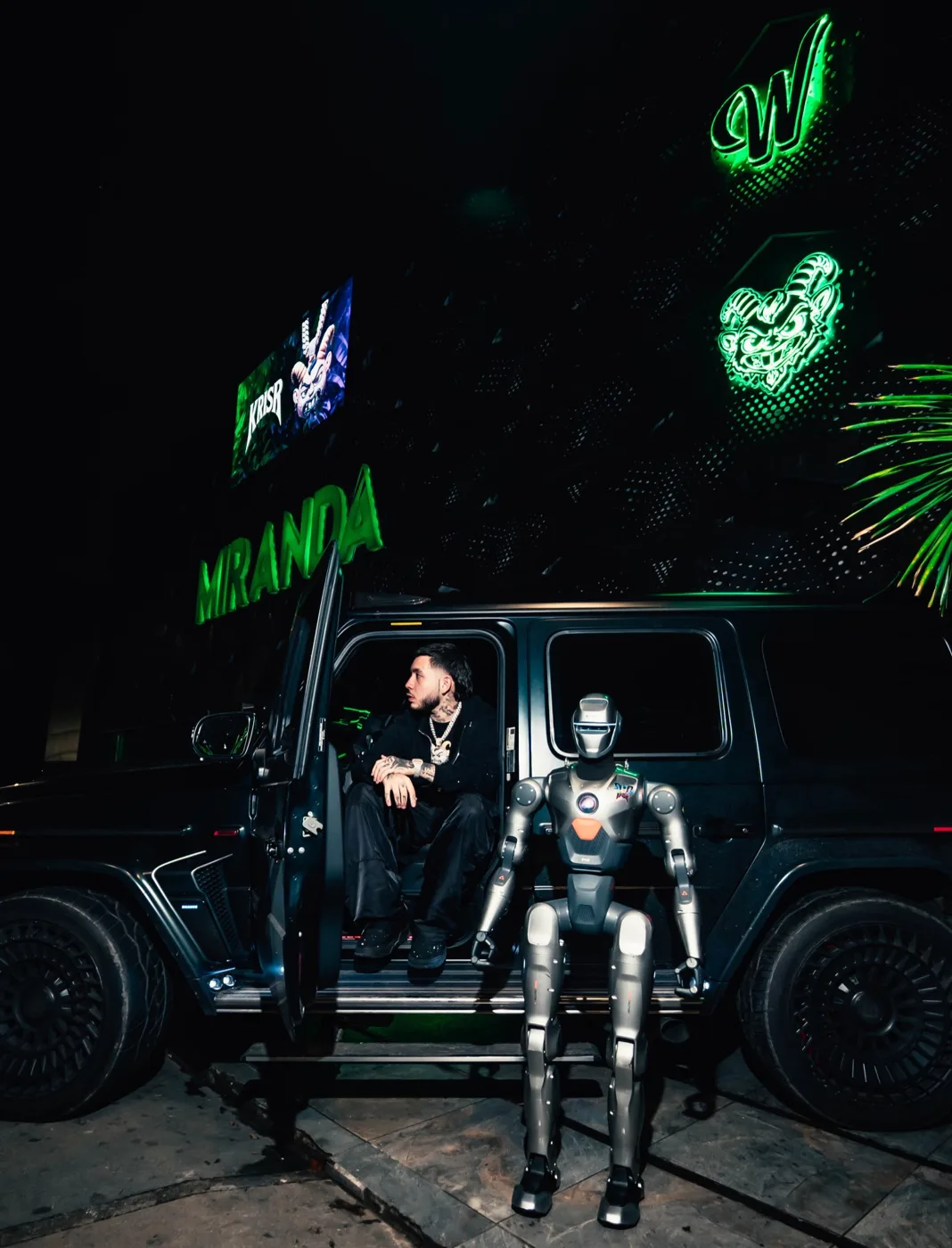 Kris R con uno de los robots de Miranda frente a la fachada con el logo en neón